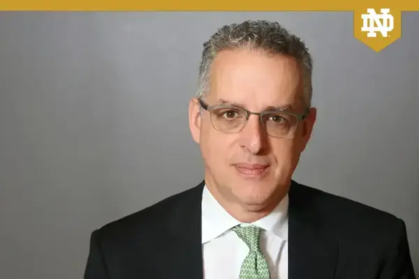 A man with gray curly hair and glasses wears a black suit, white shirt, and green patterned tie, looking forward.